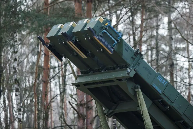 Green multi-canister missile launcher angled upward among bare winter forest trees.