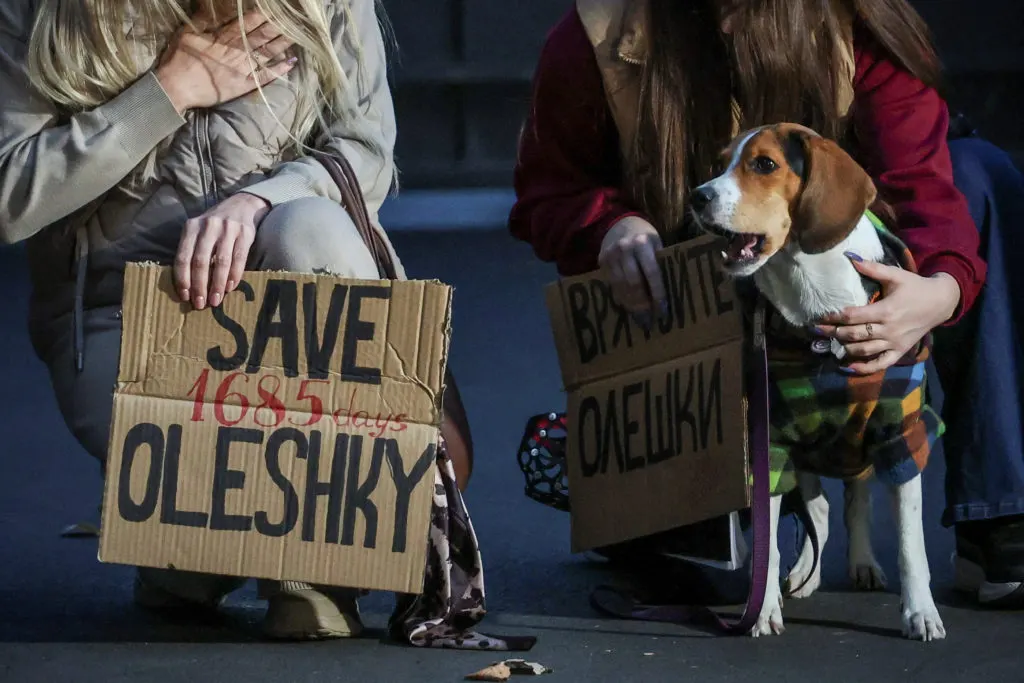 Protesters kneel with cardboard signs reading 'Save Oleshky' and a dog in a coat beside them