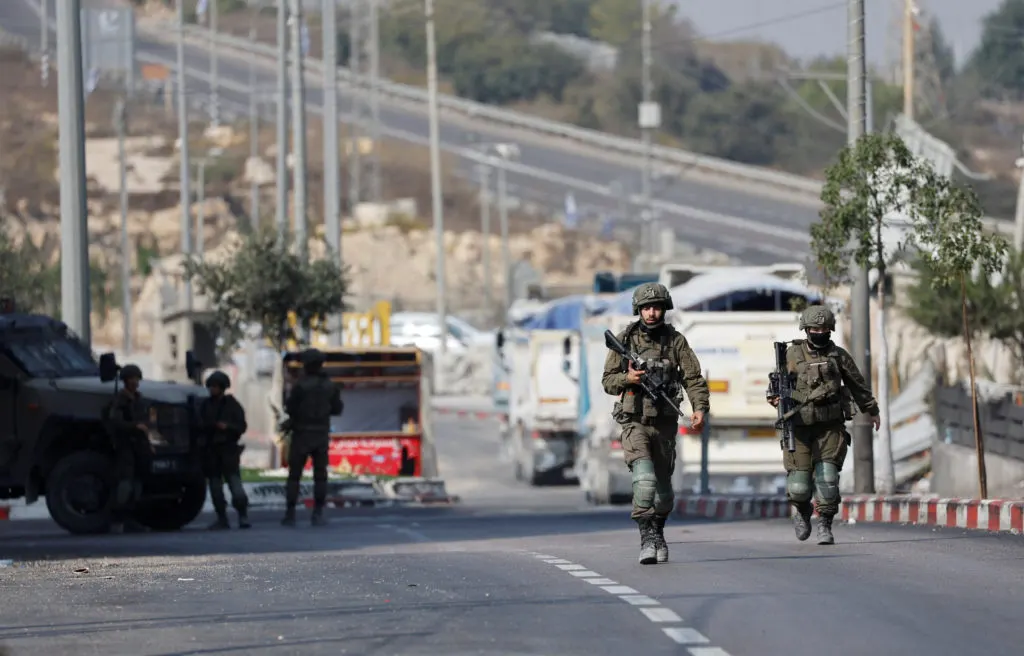 Armed soldiers in camouflage patrol a road near military vehicles and parked trucks at a checkpoint.