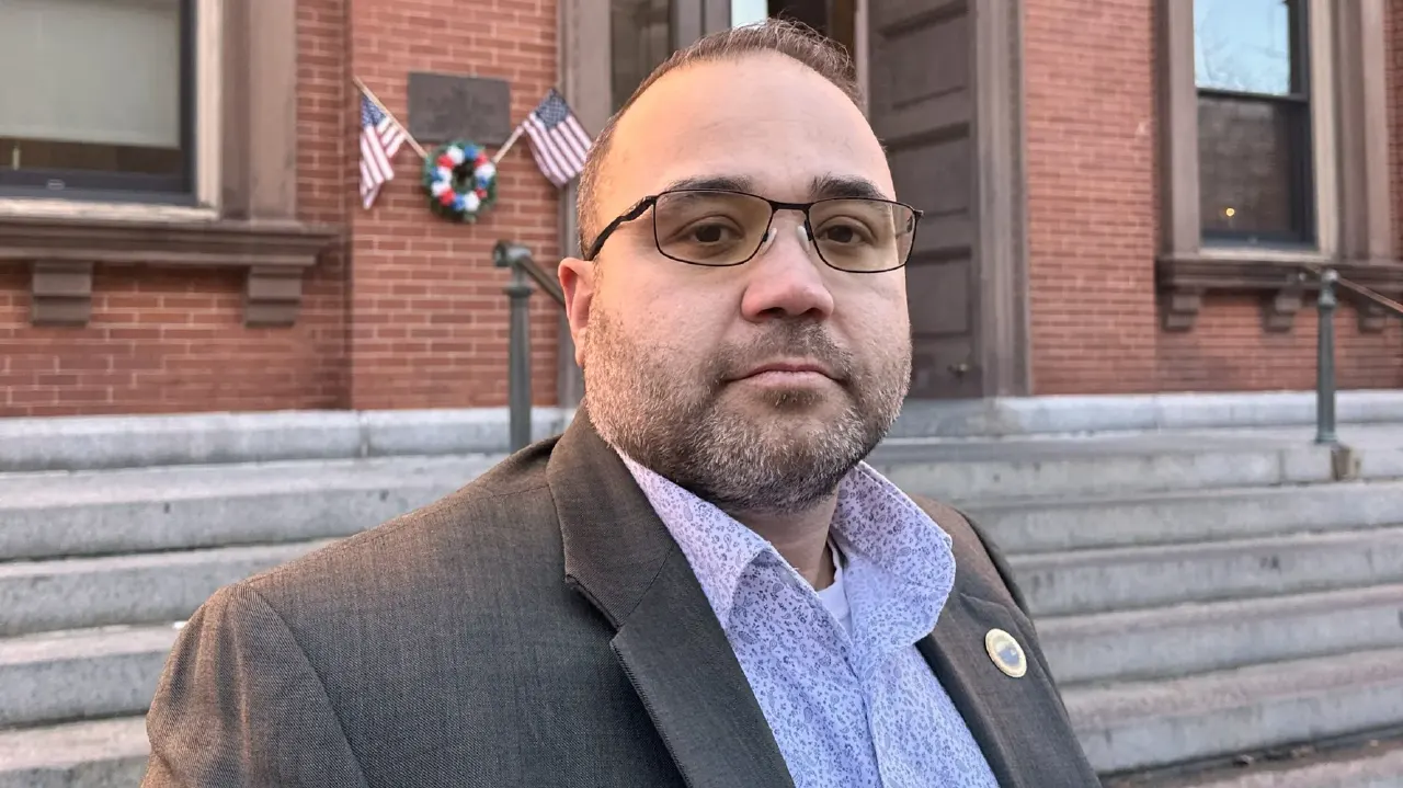 Bearded man in glasses and blazer before a brick building with American flags and a wreath.