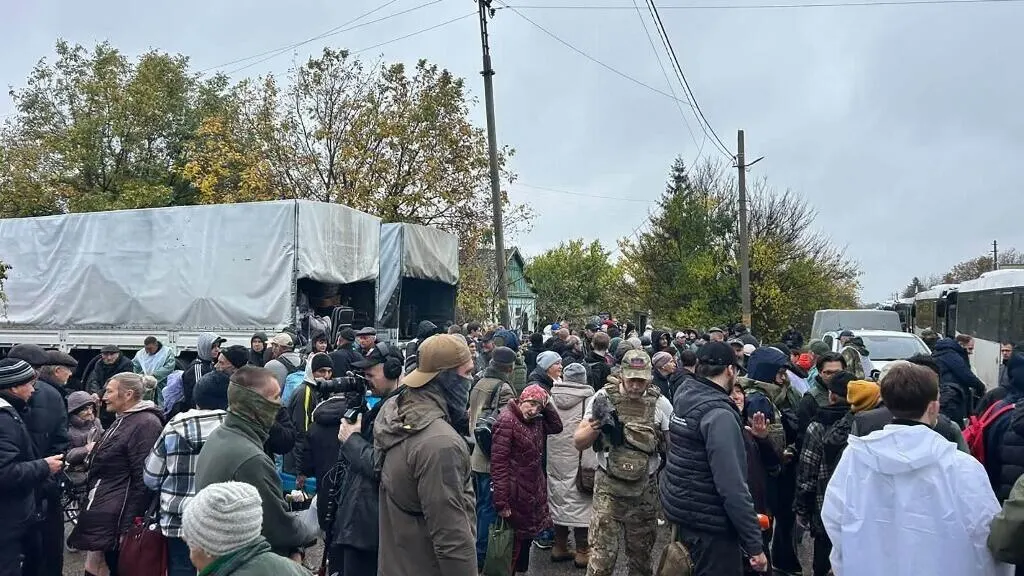 Large crowd of civilians with bags gathered beside covered trucks and buses on a rural road, some in military gear.