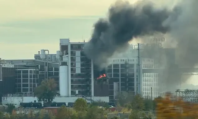 Thick black smoke and flames rising from a large industrial plant with Quaker sign visible.
