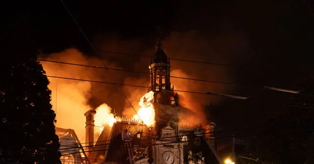 Night photo of a historic building with clock tower engulfed in flames and smoke.