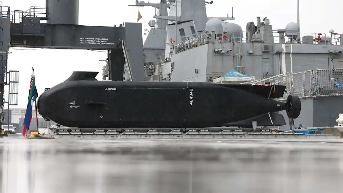 Black submarine docked at naval facility with military vessel in background.
