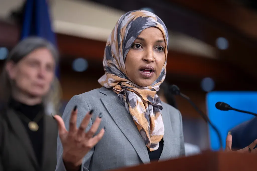 Woman in grey blazer and patterned headscarf gesturing while speaking at a press conference podium