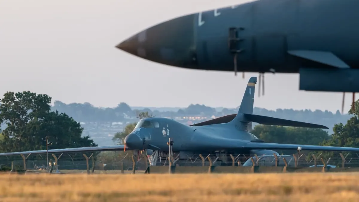 Military bomber aircraft on airfield tarmac behind perimeter fence, with another aircraft nose in foreground.