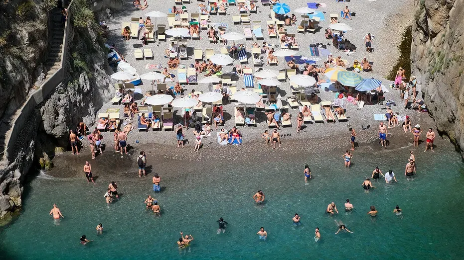 Aerial view of crowded beach with umbrellas and swimmers in turquoise water between cliffs.