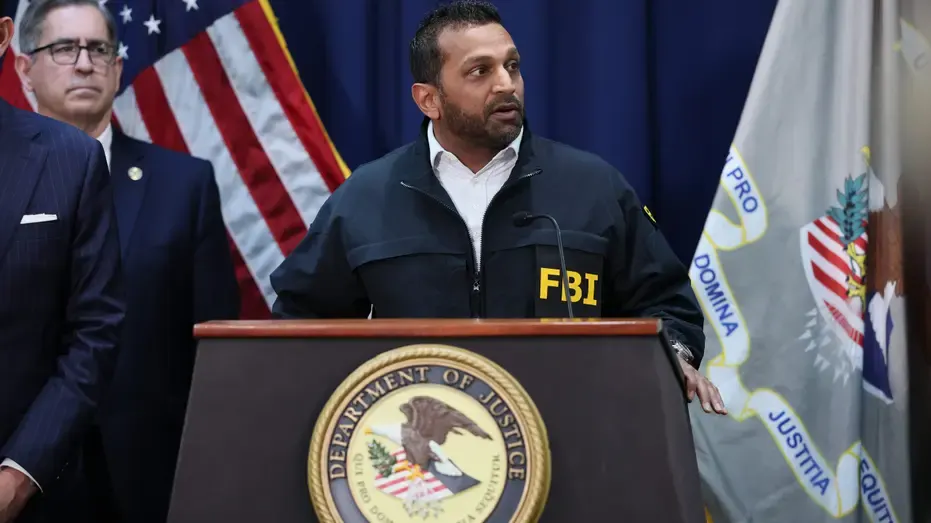 Man in FBI jacket speaking at Department of Justice podium with flags behind him.