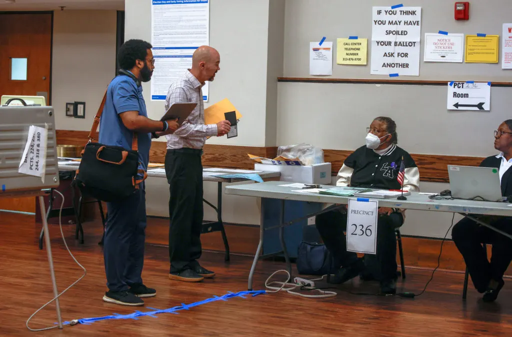 Two men stand at a polling station check-in table marked Precinct 236 while poll workers sit nearby.