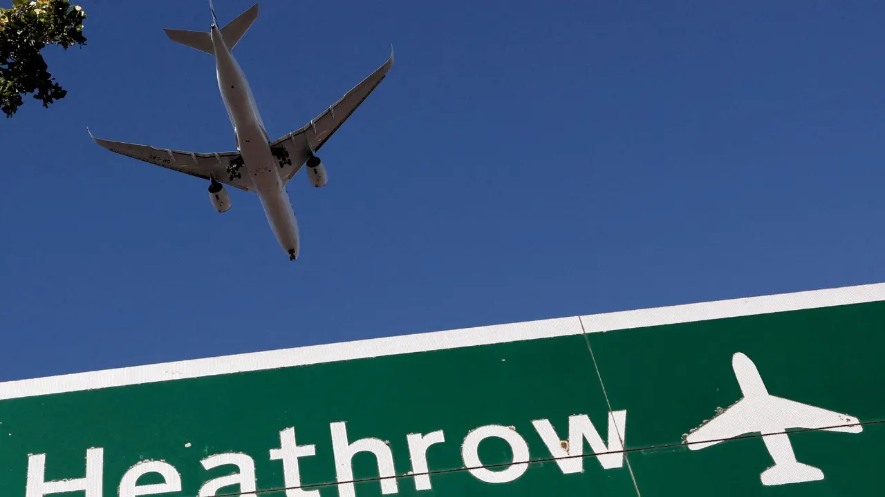 Passenger jet descending over green Heathrow road sign with airplane symbol against blue sky.