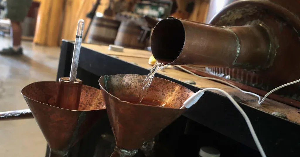 Copper still spouting clear liquid into copper cups with a thermometer, in a distillery setting.