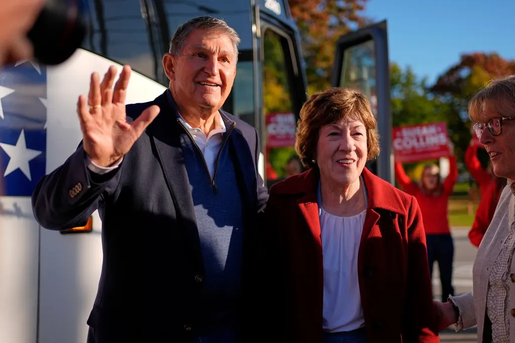 Two smiling politicians campaign outdoors, one waving, beside a bus with red campaign signs.