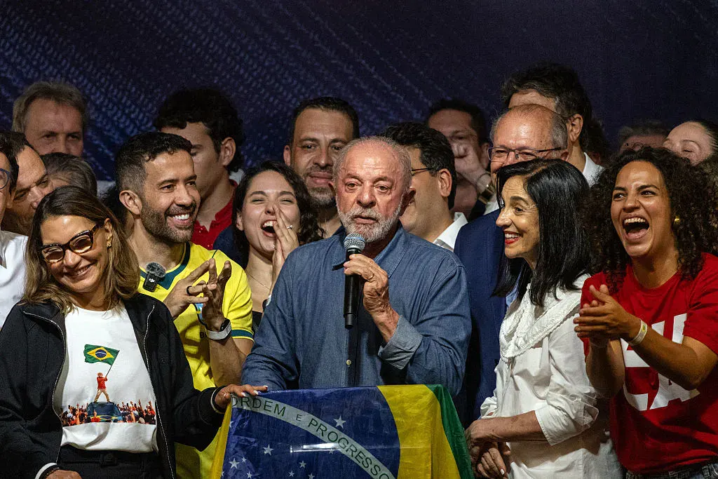 Older man speaks at a podium draped in Brazilian flag, smiling supporters gathered around him.