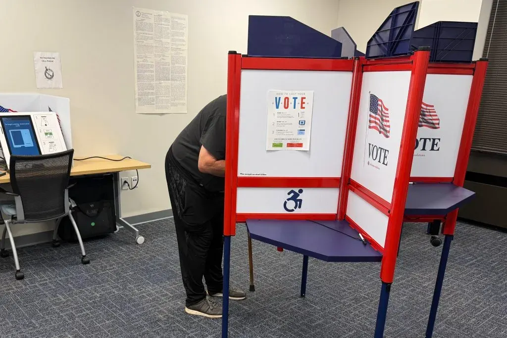 A person with face hidden votes in a red-and-white booth labeled VOTE with American flags.