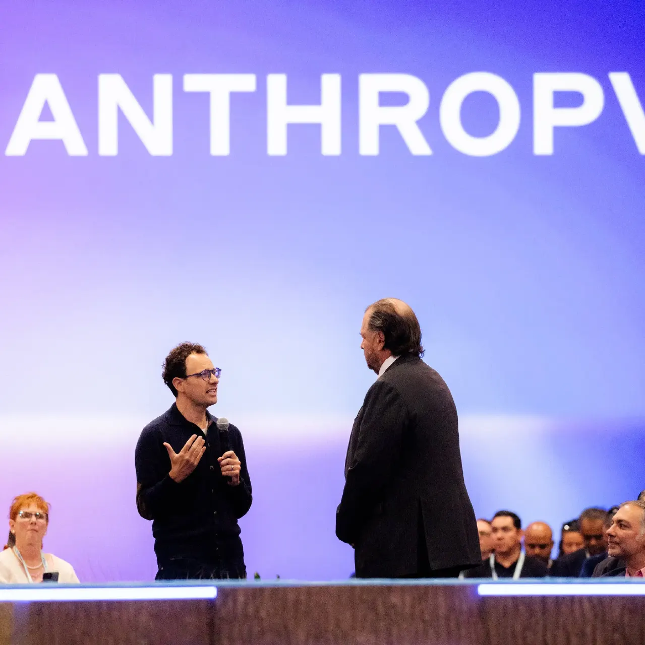 Two men talking on a stage before a large screen displaying the word ANTHROP
