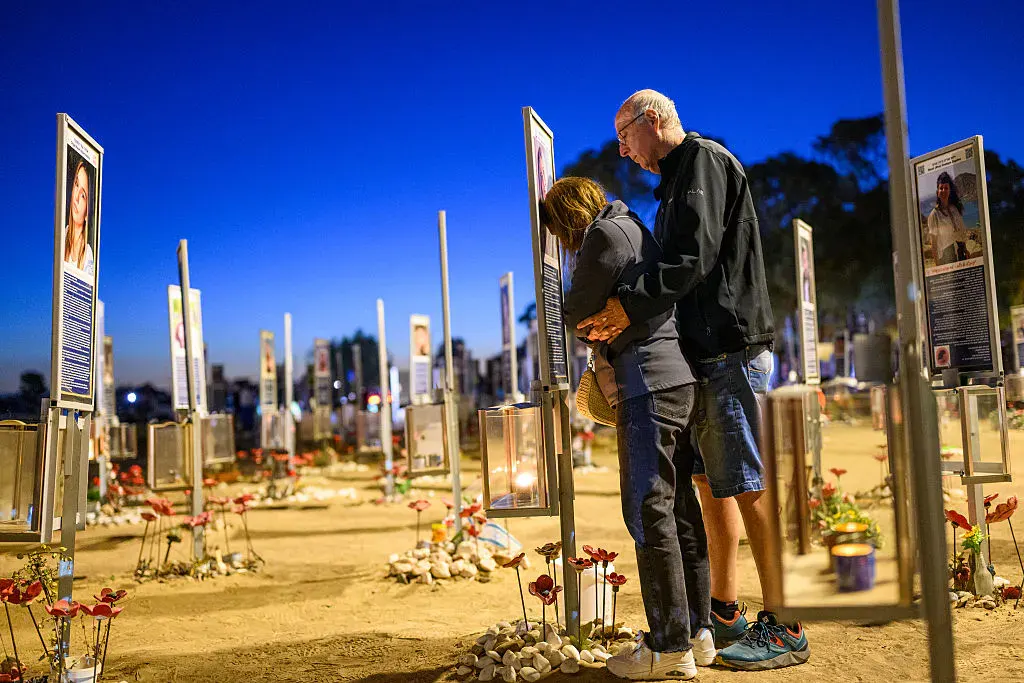 Two people stand in memorial cemetery with portraits and flowers honoring victims.