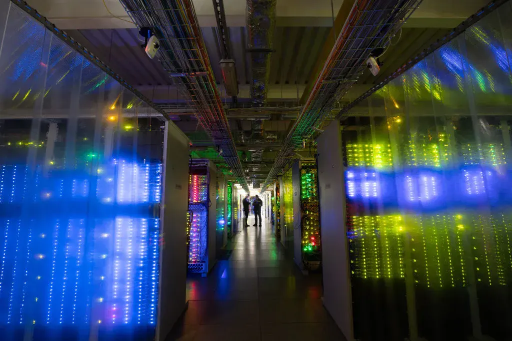 Server racks with glowing blue and green lights in a data center corridor, two people far away.