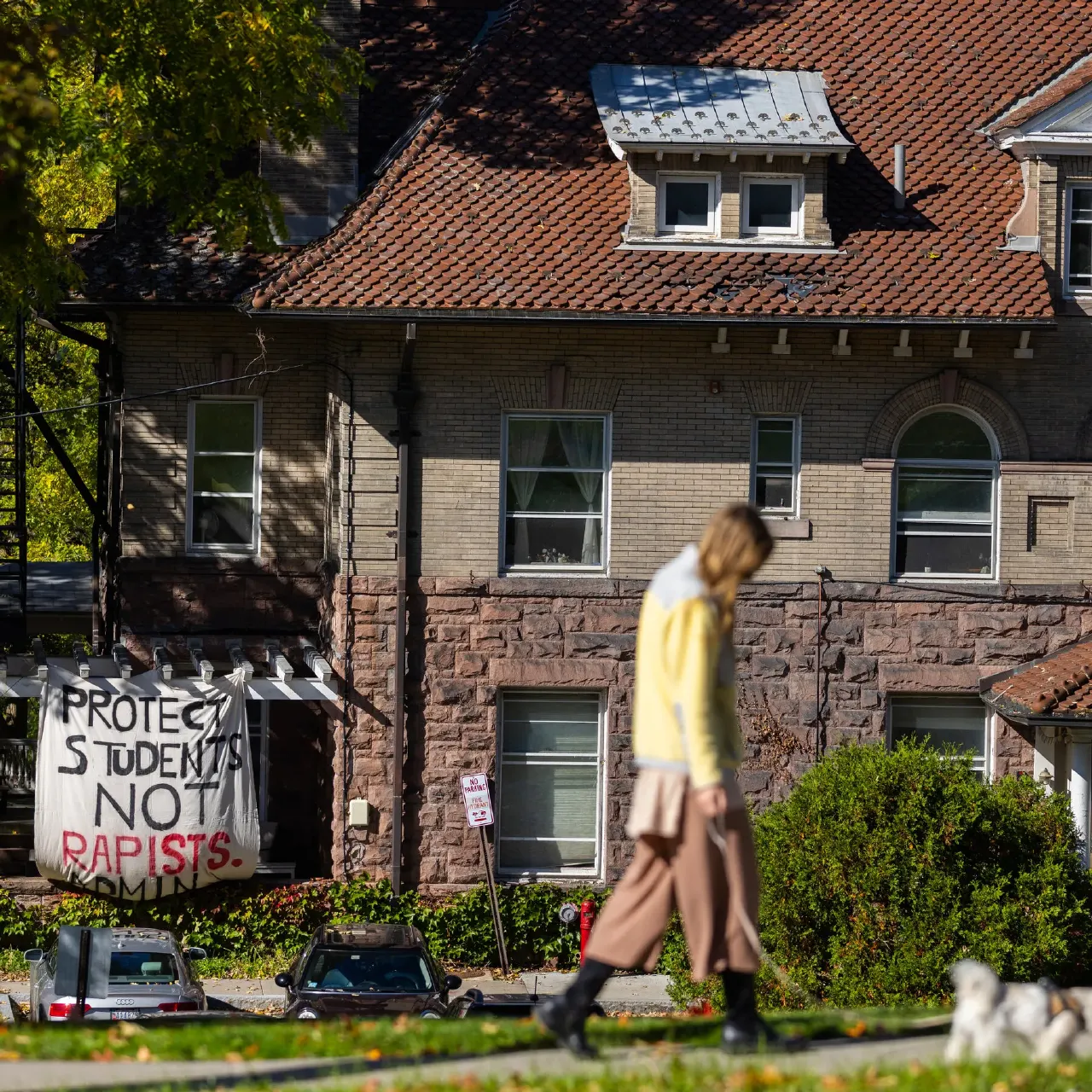 Brick fraternity house with hanging banner reading protest message about students and sexual assault, pedestrian walking