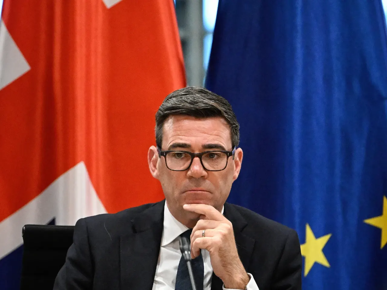 Man in glasses and dark suit, hand on chin, seated before a Union Jack and EU flag.