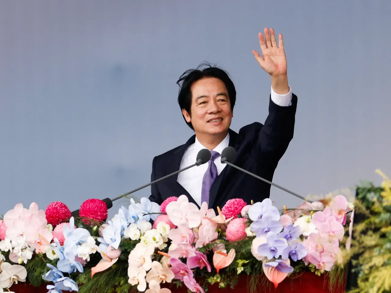 Smiling man in dark suit waving from a podium decorated with flowers, microphones in front.