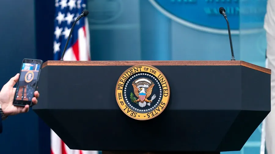 White House press briefing podium with presidential seal, flanked by American flag and microphones