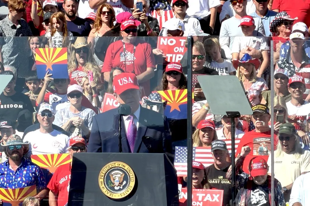Speaker at a podium with presidential seal before a crowd of supporters in red caps and Arizona flags.