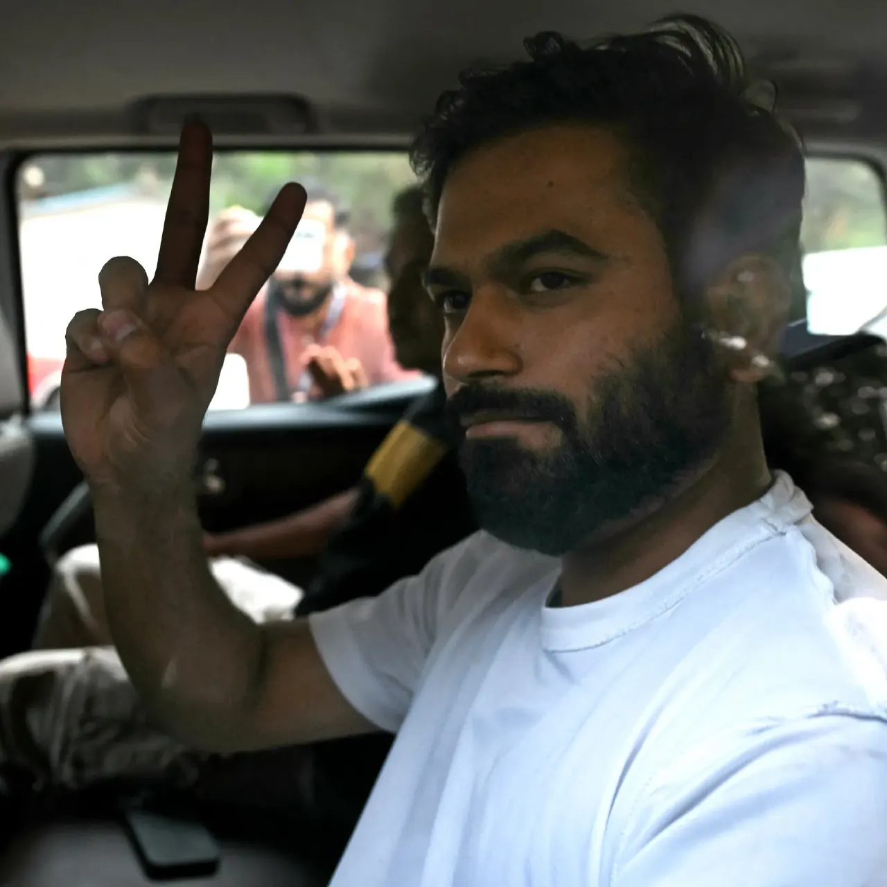 Bearded man in white shirt flashing a V sign from inside a car window