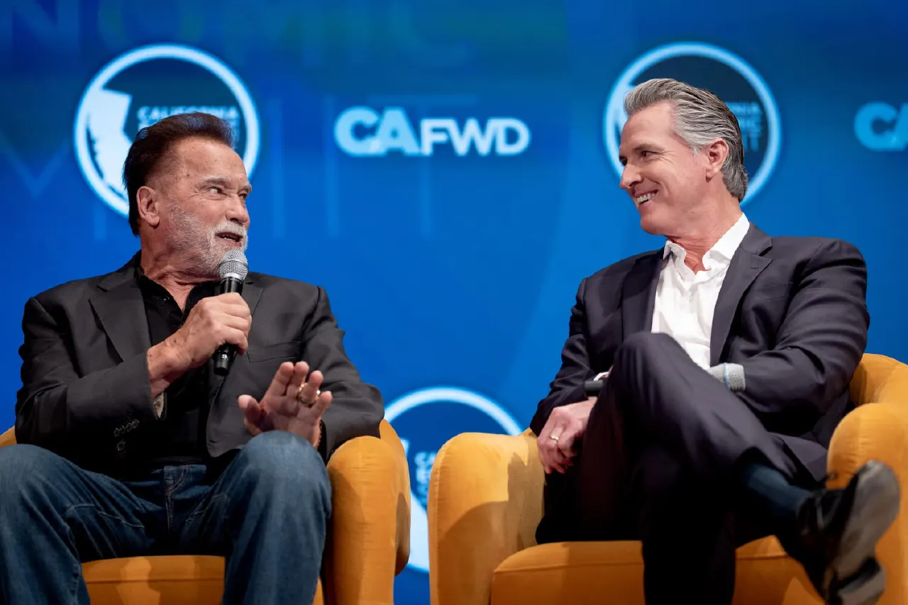 Two men seated on gold chairs in conversation on stage at a blue-backdropped summit event.