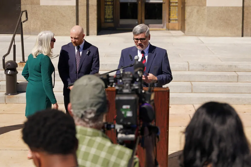 Three people speak at a outdoor podium on a courthouse plaza while camera operators film.
