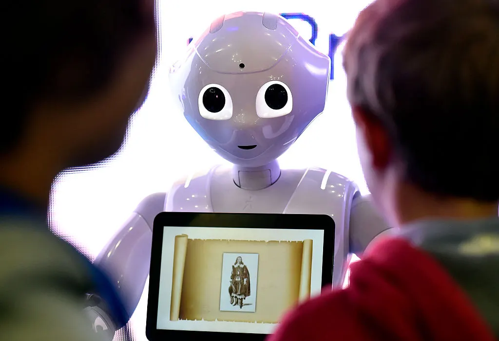 White humanoid robot with large eyes beside a tablet showing a scroll and figure, viewed by two people.
