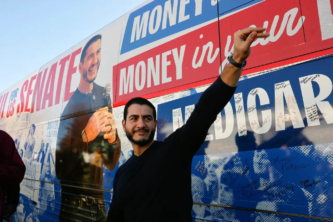 Smiling bearded man raising arm before a campaign bus with 'Senate', 'Money in your', and 'Medicare' slogans