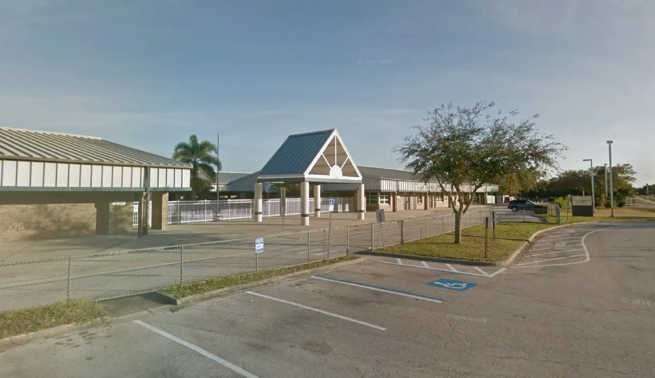 Street-level view of a single-story school building with a gabled entrance canopy, fence, and empty parking lot.