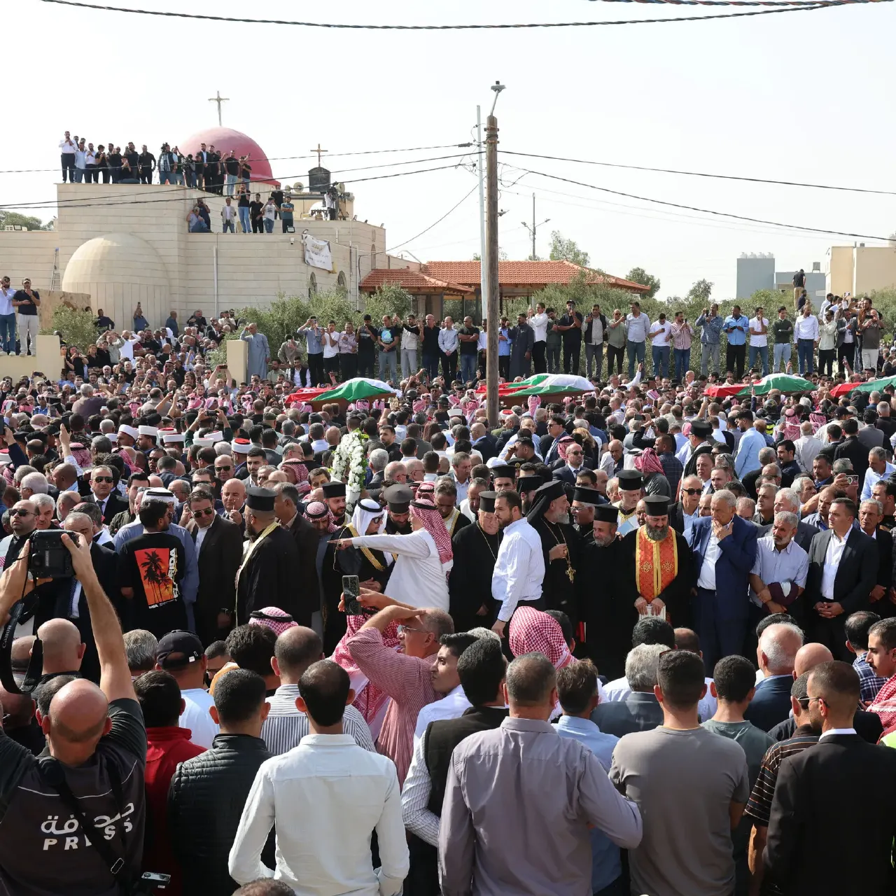 Large crowd of mourners in black and clerical robes gathered outside a church with a red dome.