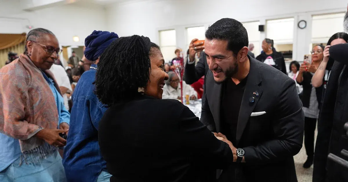 A smiling man in a dark suit greets a smiling woman at an indoor community gathering, people around them.