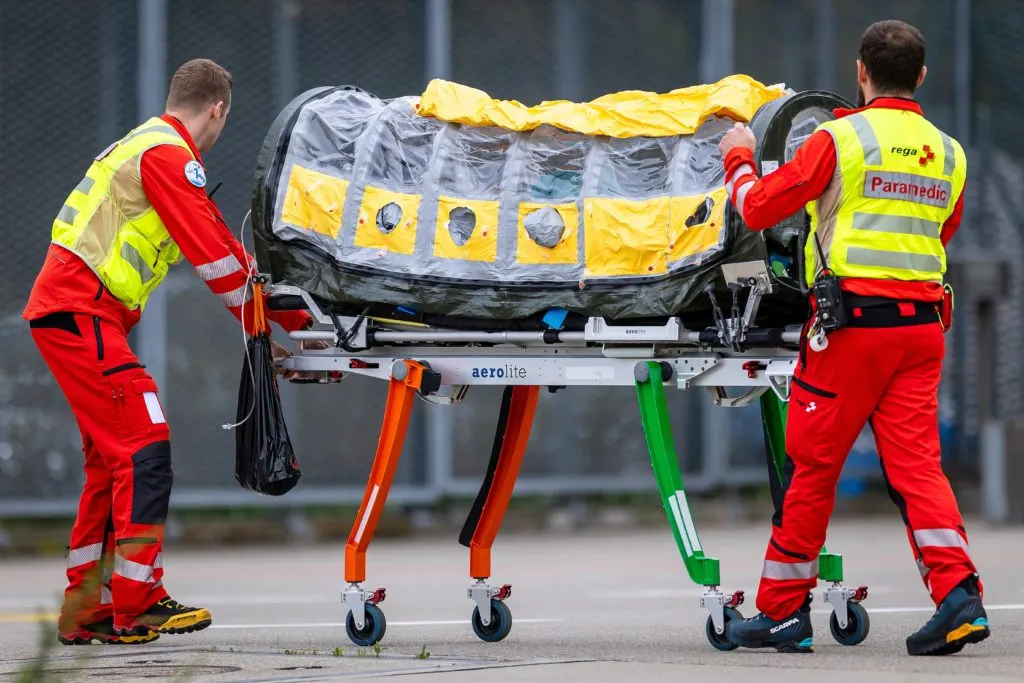 Paramedics in red and yellow uniforms wheel an enclosed isolation stretcher across a tarmac.