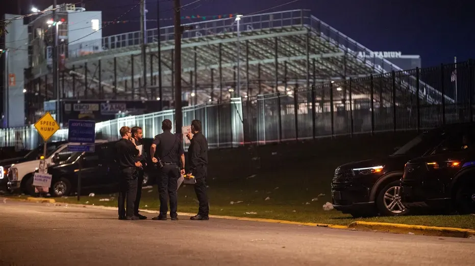 Uniformed officers talk on a dark street near a stadium fence, with parked police SUVs and litter on the grass.