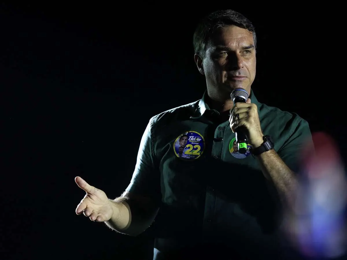 Man in dark shirt with campaign buttons speaking into microphone against dark background