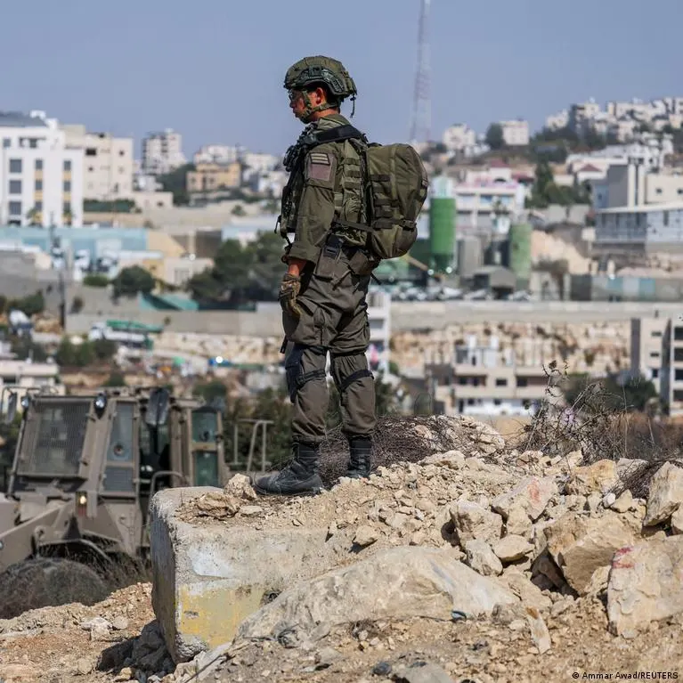 Soldier in helmet and fatigues stands on rubble beside a military vehicle, overlooking a hillside town.