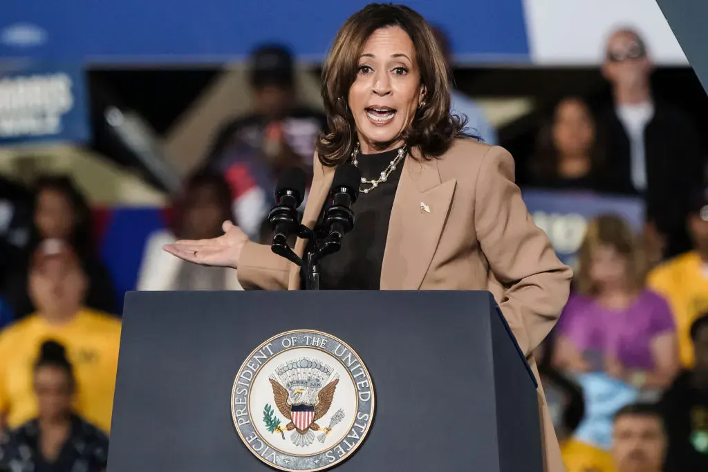 Woman in tan blazer gesturing while speaking at a podium bearing the Presidential seal.