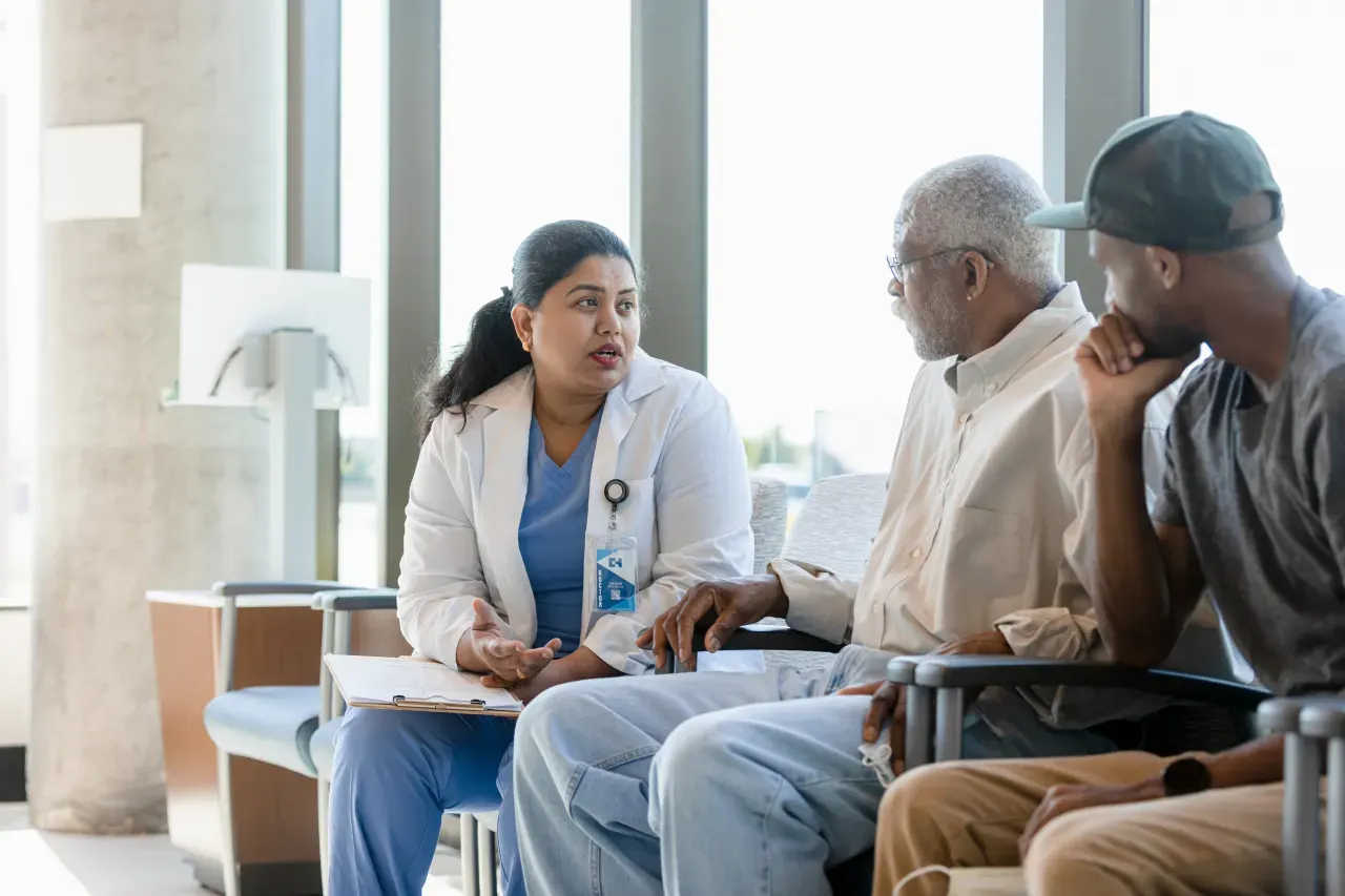 Female healthcare worker in scrubs and white coat speaks with two older patients in a waiting room