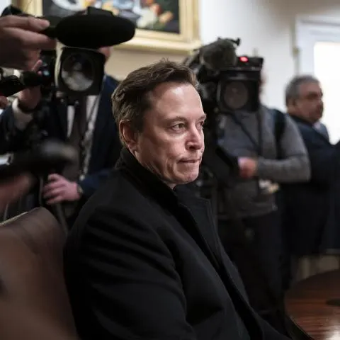 A man in a dark coat sits while news camera crews and reporters surround him in a room.