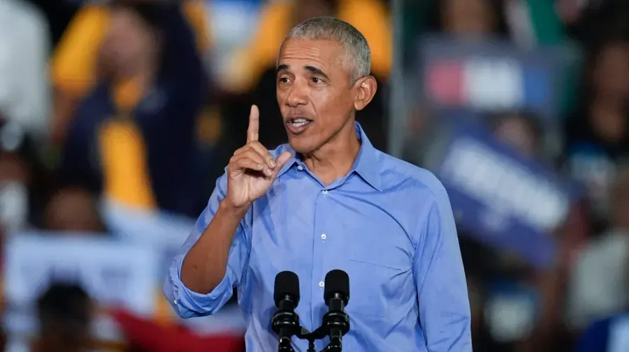 Man in blue shirt raising finger while speaking at a podium with blurred crowd behind.