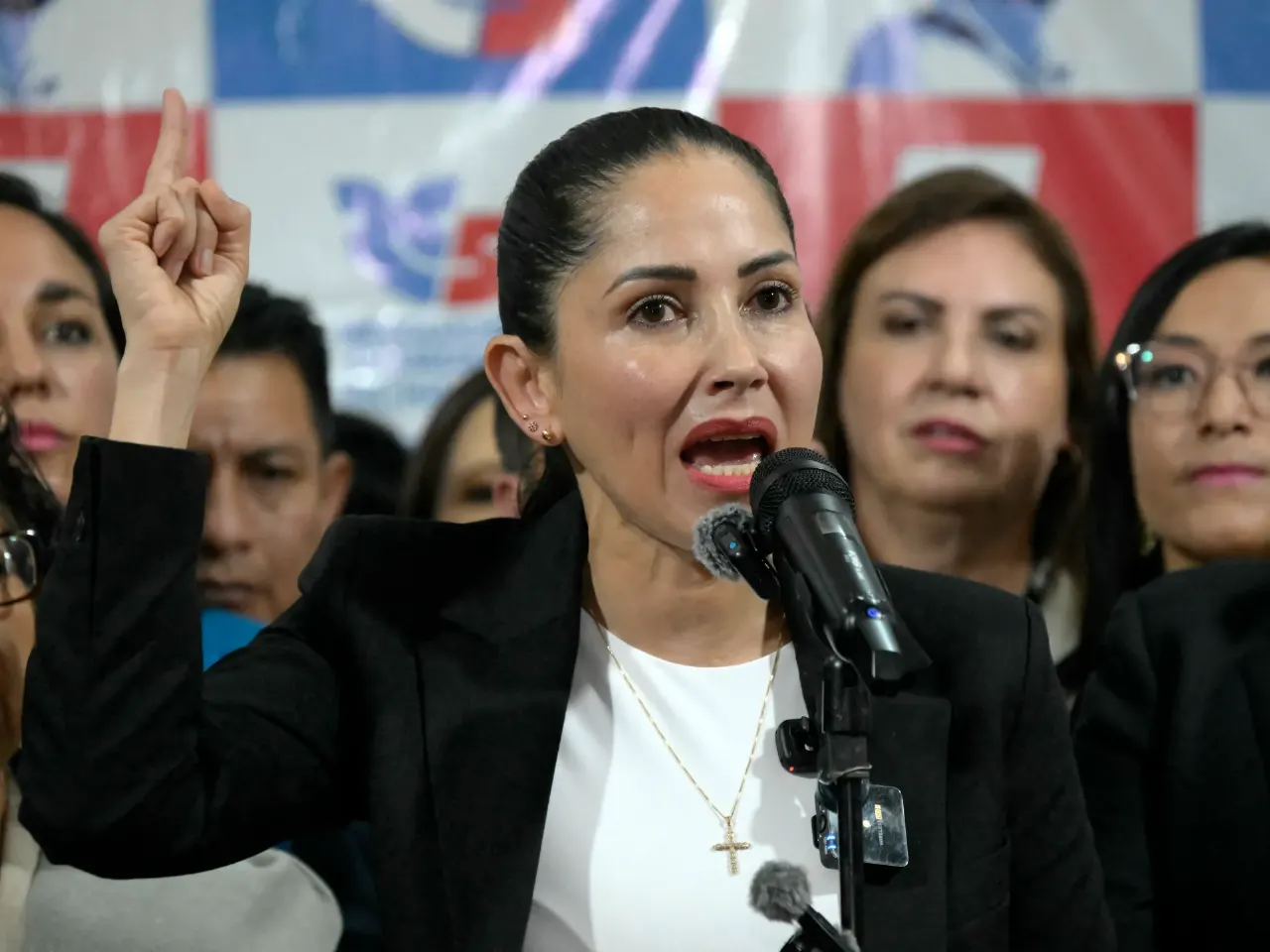Woman in black blazer speaks at microphone, raising index finger, before blue-and-red party backdrop with supporters.