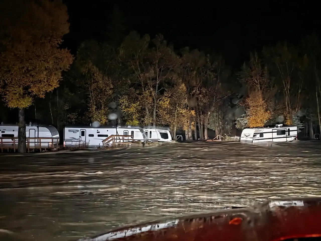 Night photo of fast brown floodwaters surrounding several travel trailers among trees.