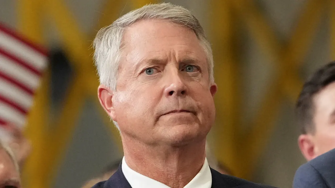 Older man with gray hair in dark suit and white shirt, with American flag blurred behind him.