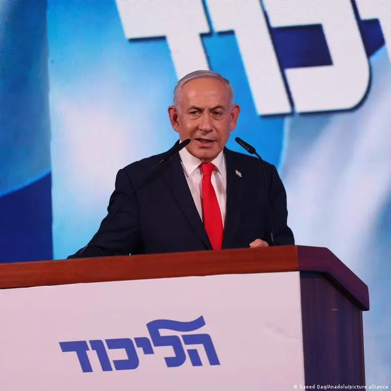 A man in a dark suit and red tie speaks at a podium bearing the Likud party logo.