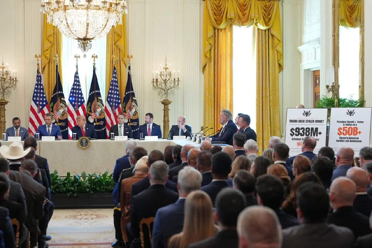 Officials seated at a formal White House roundtable with American flags, facing a seated audience and signage.