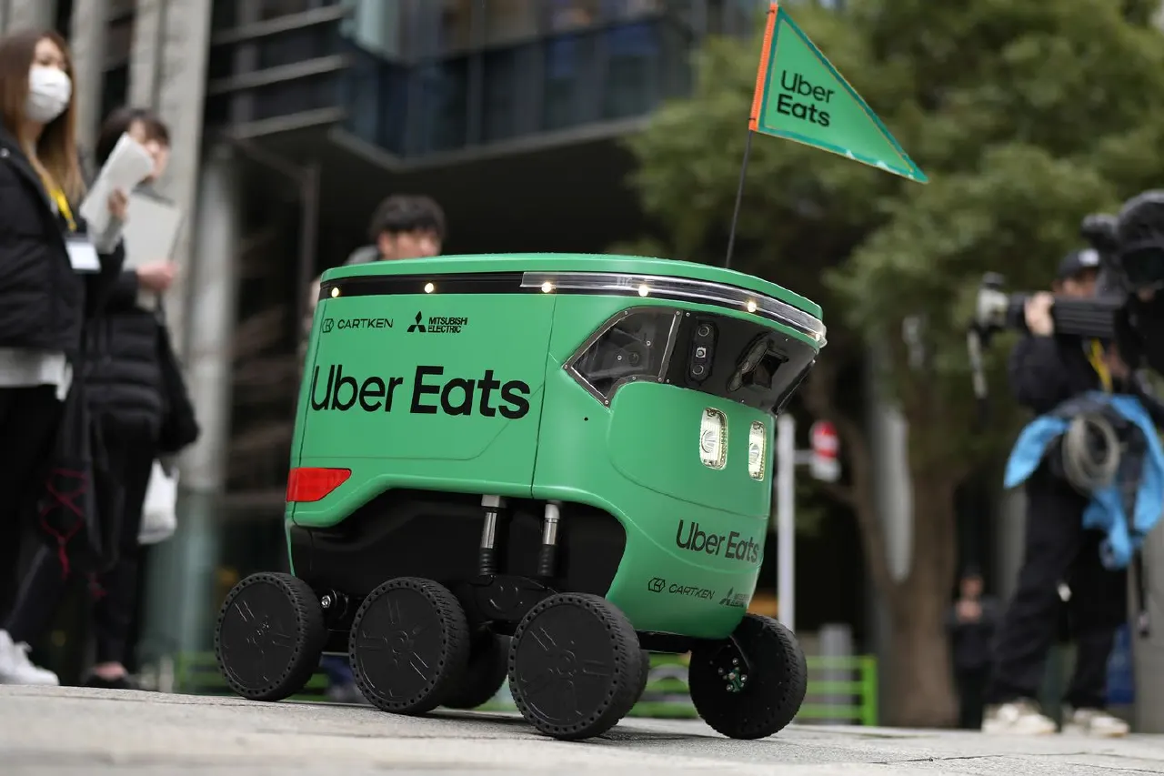Green Uber Eats delivery robot on a city street with pedestrians and a photographer nearby.