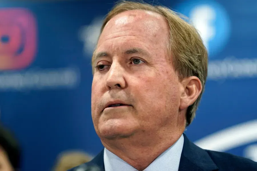 Middle-aged man in suit speaking at a press event before blue backdrop with blurred logos.
