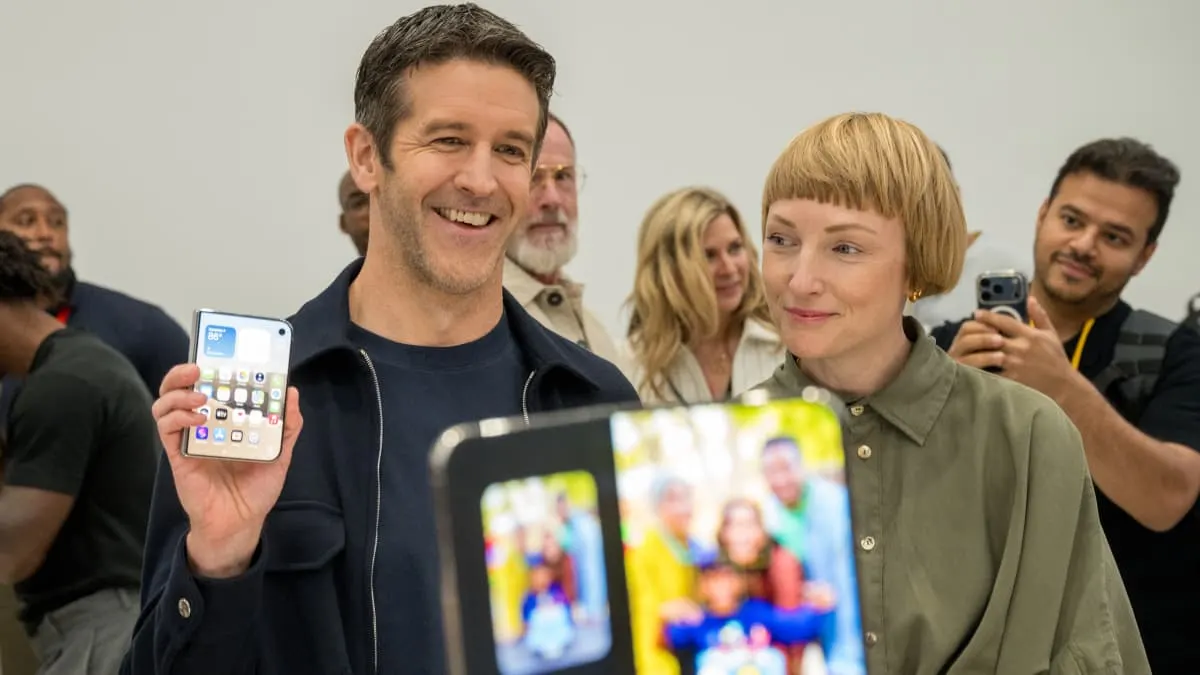Smiling man holds up foldable phone as woman and onlookers watch at event.
